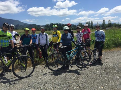 A group of riders pause for a group photo with field, hills, and clouds in background.