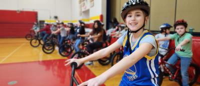 A student wearing a helmet smiles as they get ready to ride a bike during gym class.