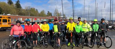 A group of riders in colorful rain jackets in yellow, green, and pink, stand in front of Cascade Bicycle Club and Sandpoint Sail's sailboats.
