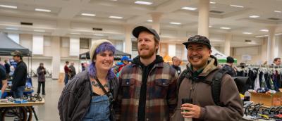 Three smiling people standing in front of vendor booths, clothing racks, and tires in a large hall with columns.