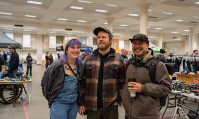 Three smiling people standing in front of vendor booths, clothing racks, and tires in a large hall with columns.