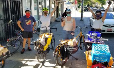 Four excited volunteer riders in helmets standing by their bikes that are loaded with deliveries to local organizations