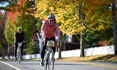 Two riders on a country road pass by rows of colorful trees and a white picket fence.