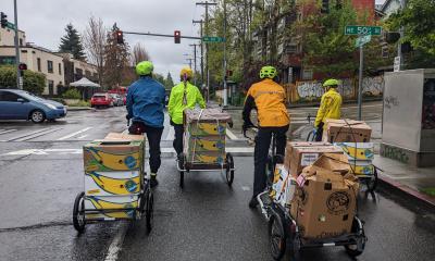 Four riders with Burley trailers stacked with banana boxes stop at an intersection.