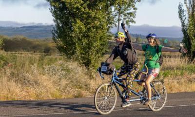 Two joyous riders on a tandem pass by low hills, sports fields, and trees. 