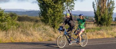 Two joyous riders on a tandem pass by low hills, sports fields, and trees. 