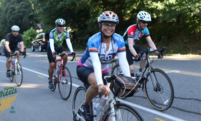 Four bicyclists riding towards and past the camera along a roadside wearing helmets and distance riding gear. The woman on the closest bike is looking at the viewer and grinning.